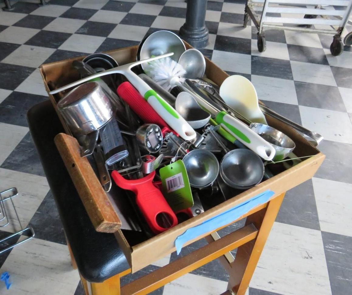 Wooden box overflowing with kitchen utensils on a wooden stool, set against a checkered floor.