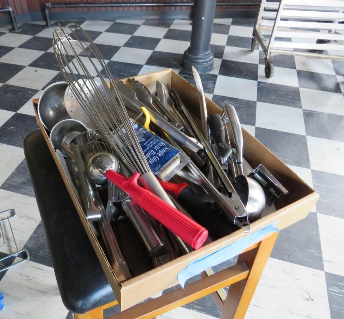 Box of assorted metal kitchen utensils on a checkered floor.
