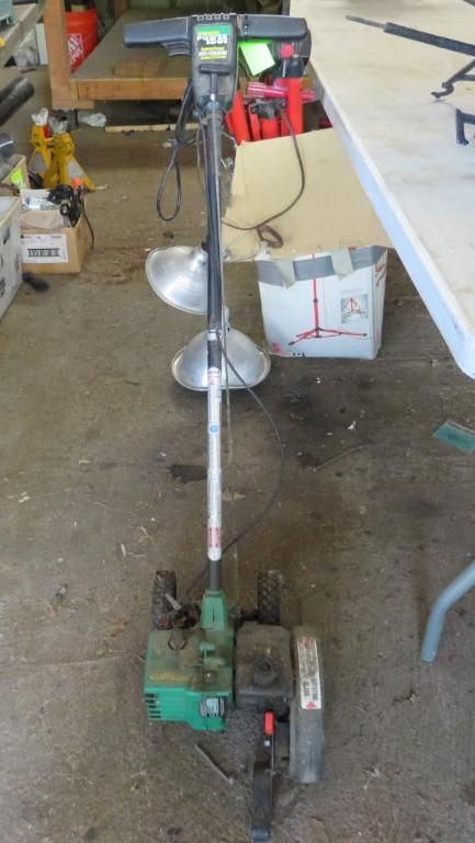 Green and silver ice auger drill with attached lights, indoors on a concrete floor.