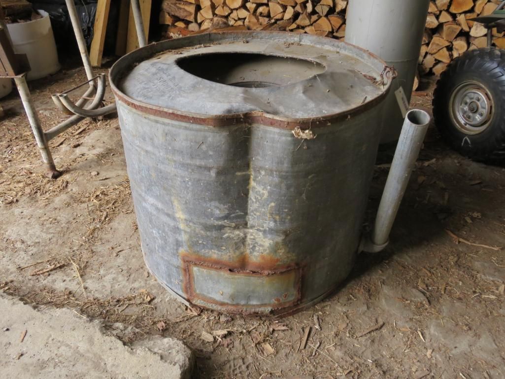 Metal barrel stove with a large hole on top, side pipe, and rectangular opening; rustic setting.