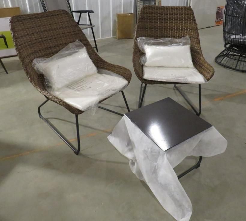 Two wicker chairs with cushions and a small table, all with protective wrapping, in a warehouse.