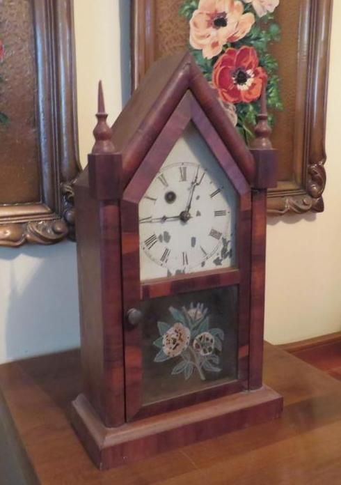 Antique wooden clock with a glass door, resting on a table in front of floral paintings.