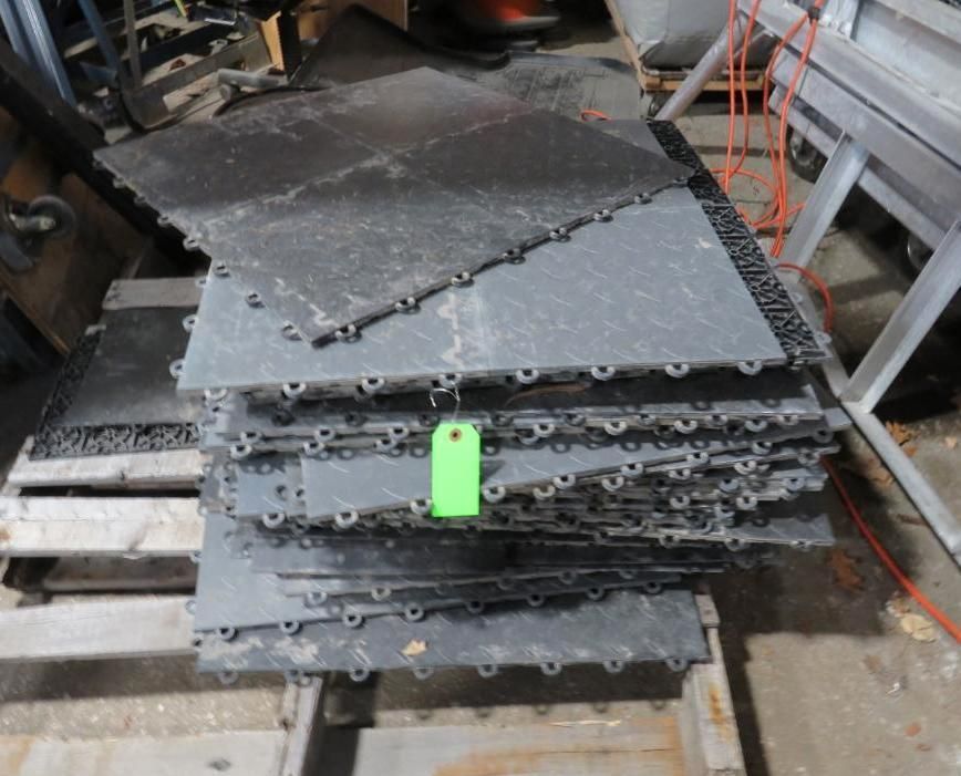 Pile of interlocking black floor tiles on a wooden pallet in a warehouse setting.