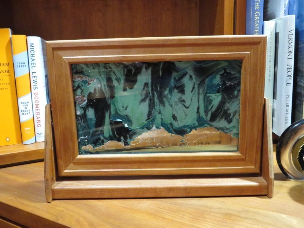 Wooden framed sand art on a shelf in front of books. Swirled blues and greens.