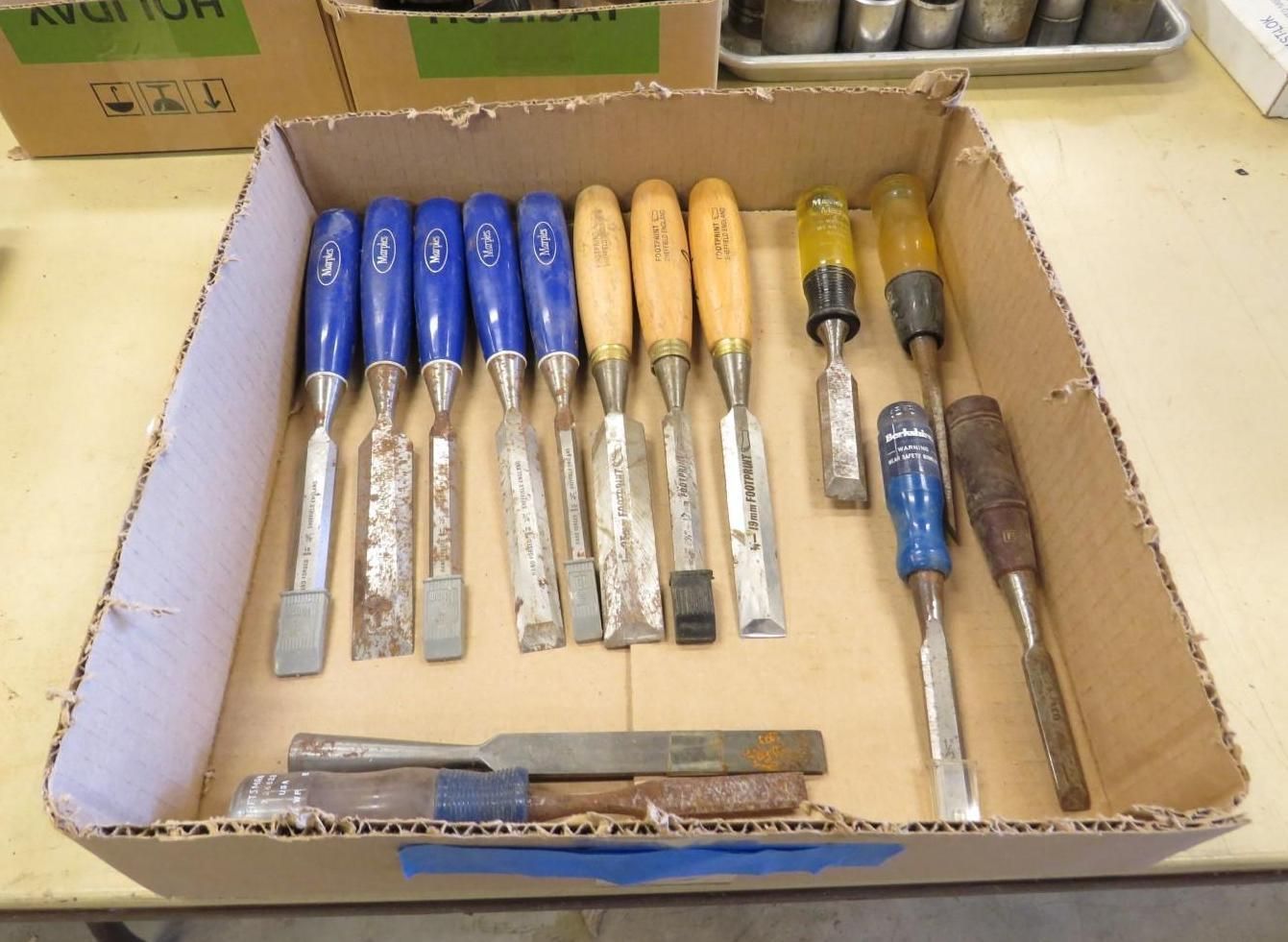 A cardboard box filled with various chisels, some with blue, yellow, and brown handles.