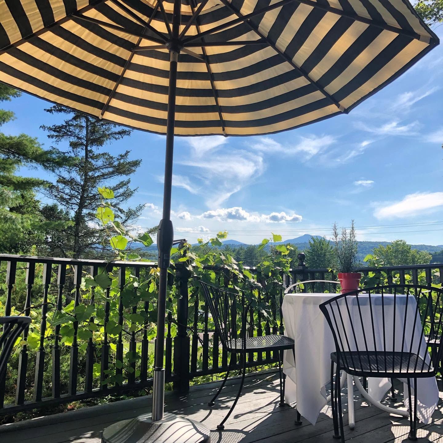 Patio with striped umbrella, table and chair, overlooking green trees and mountains under a bright blue sky