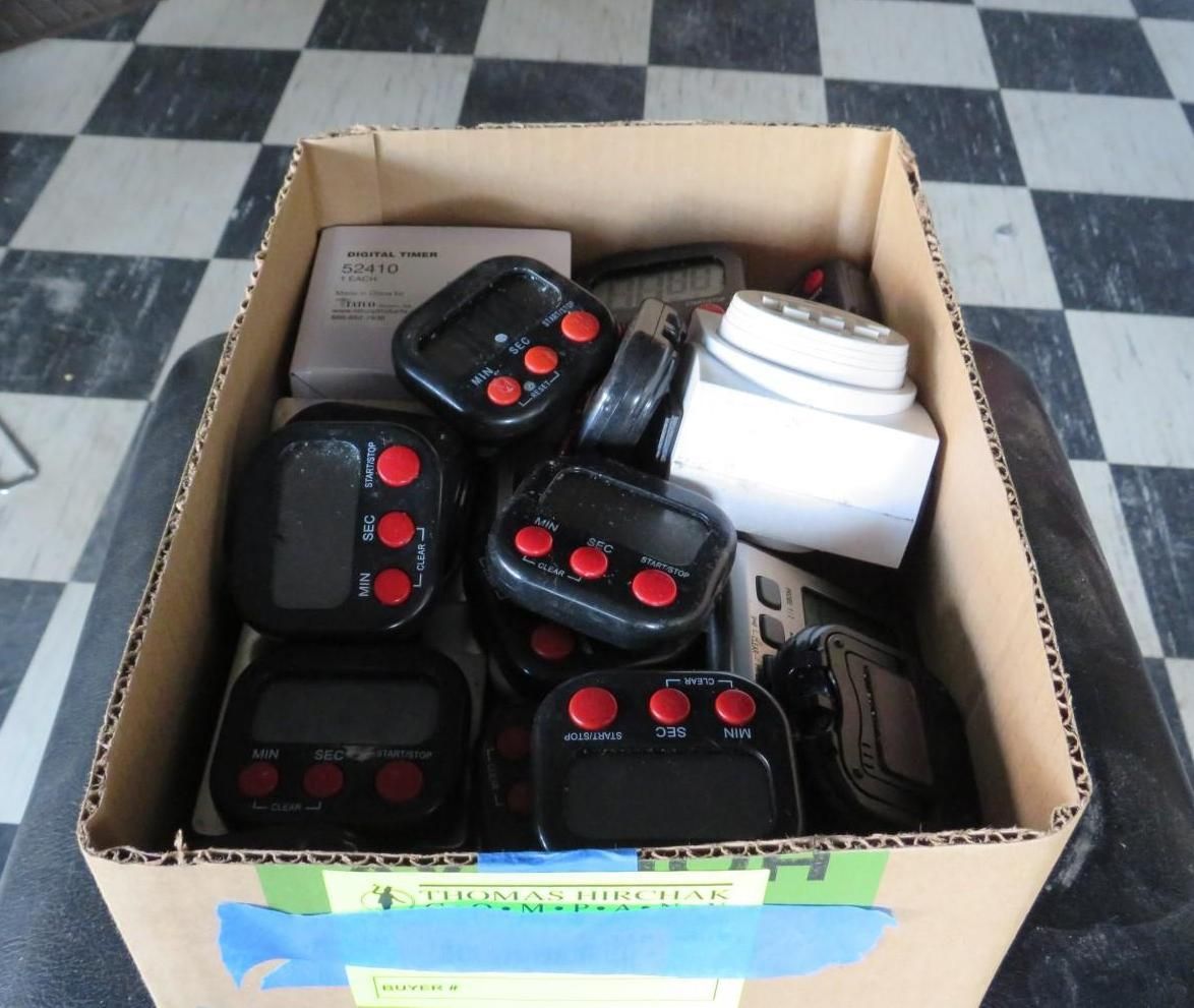 Cardboard box filled with black kitchen timers with red buttons, on a checkered floor.