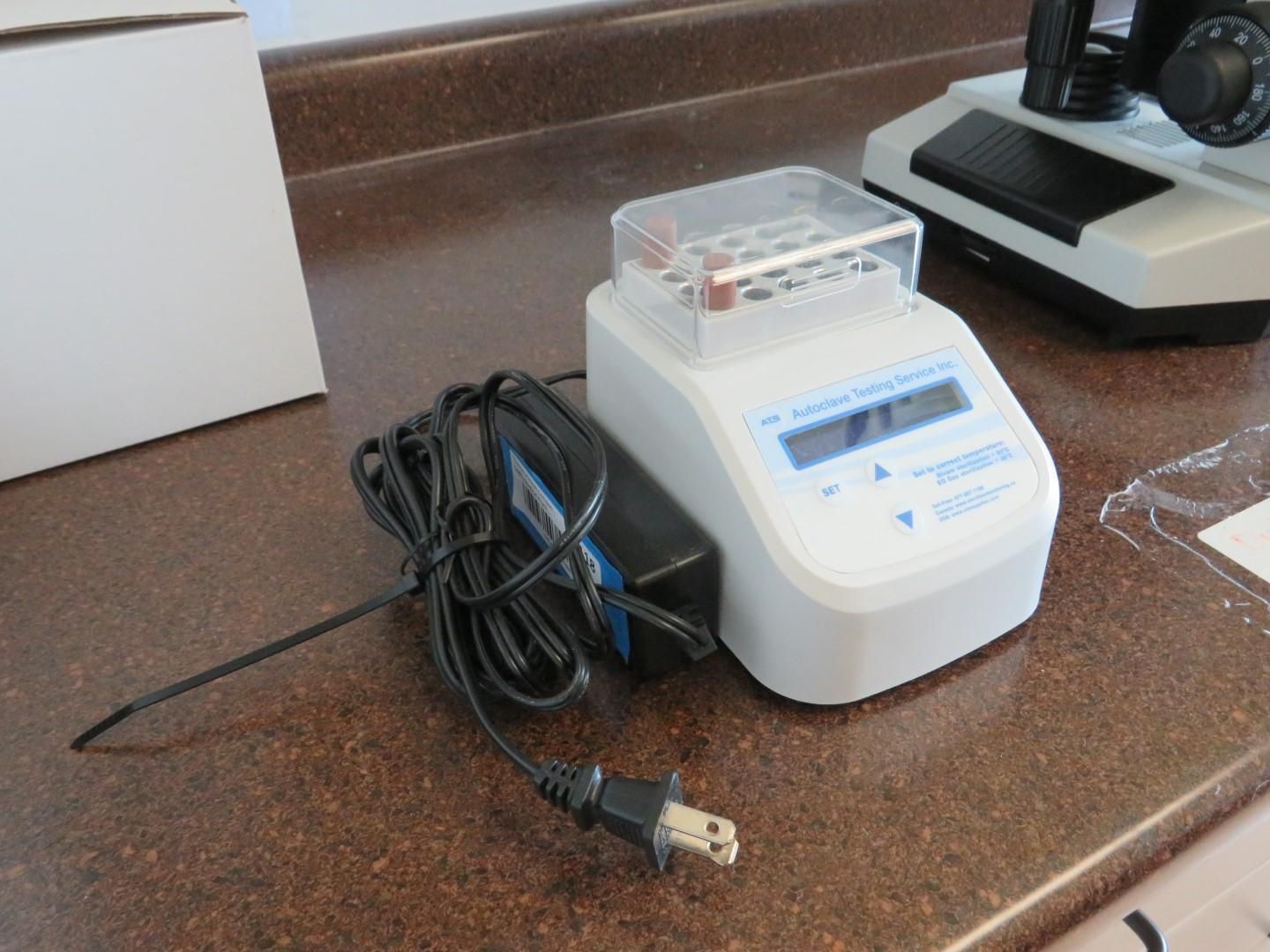 A white lab device with a clear lid, power cord, and a black power adapter, next to a microscope.