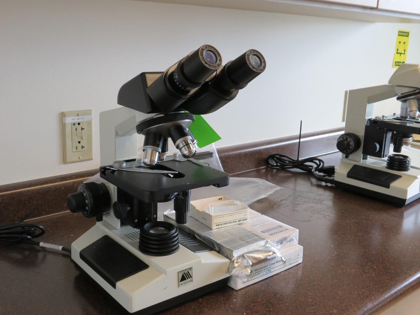 A binocular microscope with slides and another microscope on a counter in a lab setting.