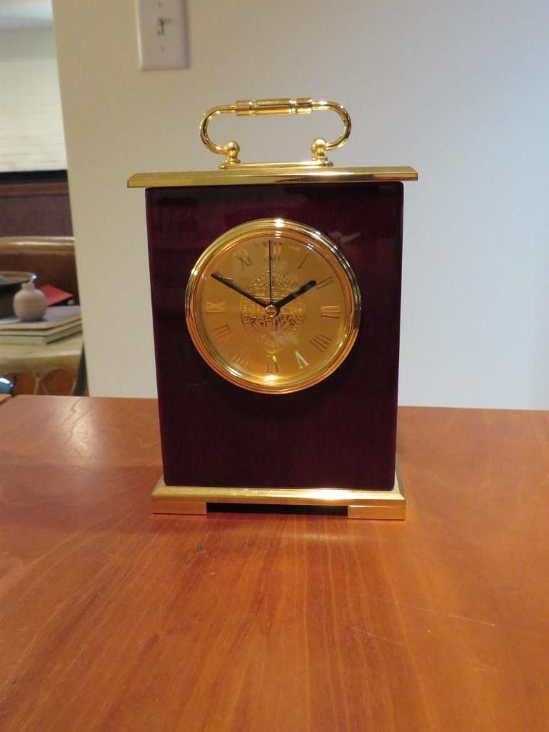 A maroon and gold clock with a handle sits on a wooden table.