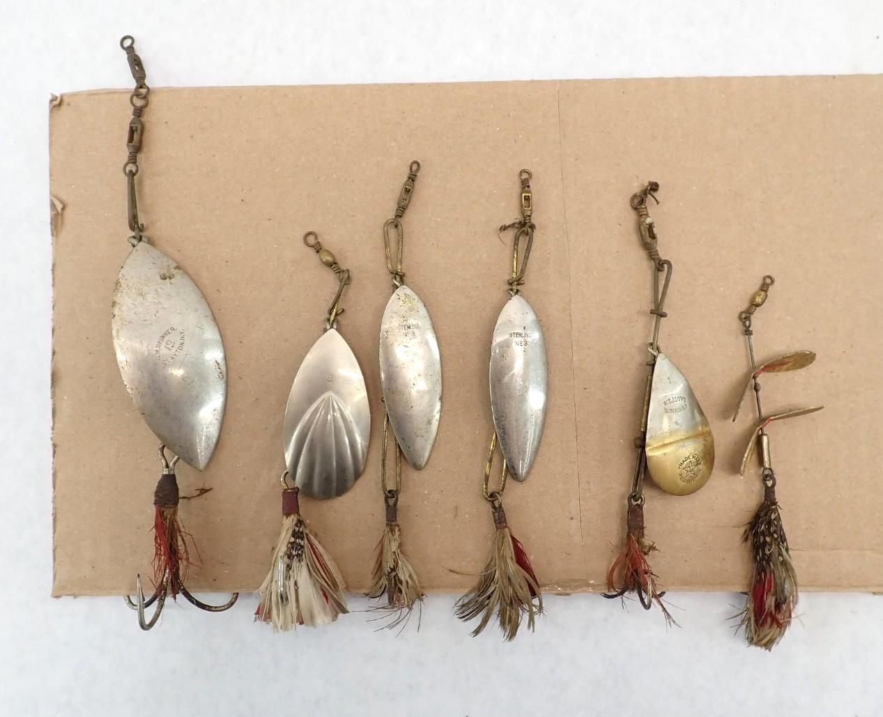 Six small fishing lures with chains and tassels, mostly silver with one gold and brown lure, on a beige board