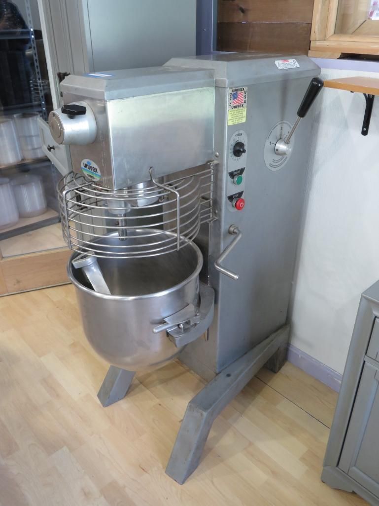 Commercial stainless steel stand mixer with a large bowl and safety cage in a kitchen setting.