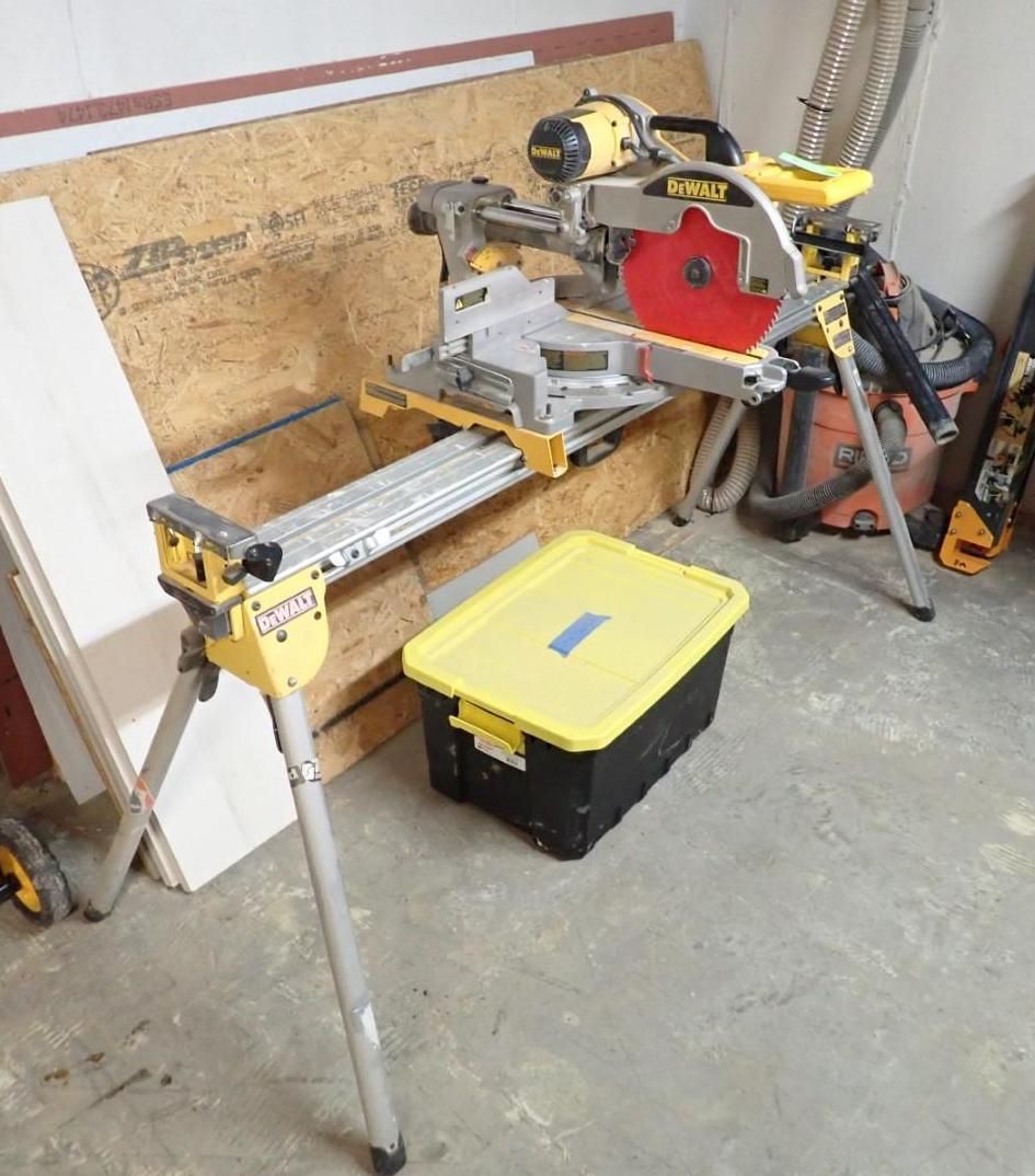 A yellow and black DeWalt miter saw on a stand, next to plywood and a storage bin in a workshop.