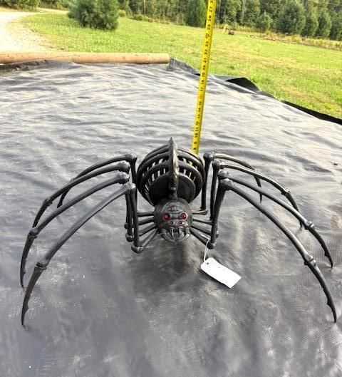 Metal spider sculpture with red eyes, sitting on black plastic, yellow measuring tape in the background. Halloween Decor.
