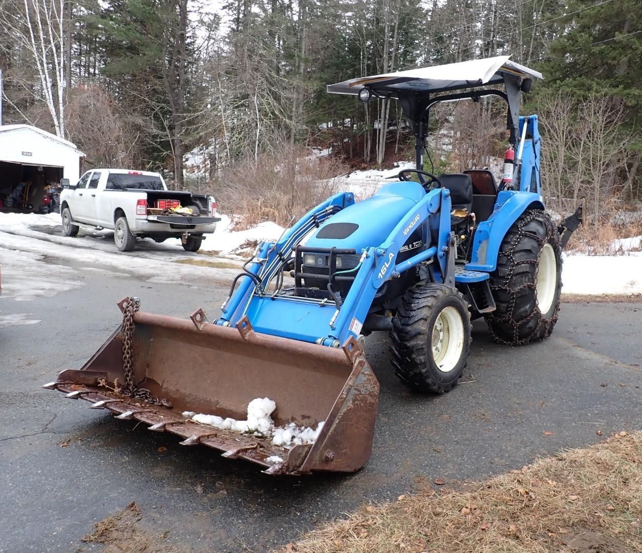 A bright blue tractor with a large front-loading bucket parked on a paved driveway near a white pickup truck.