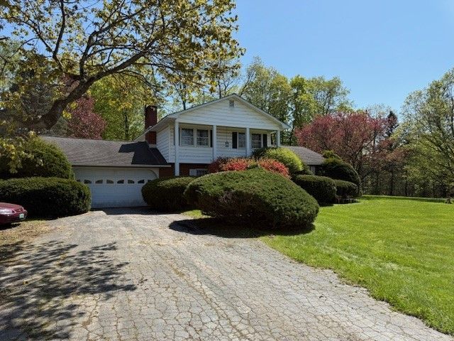 White house with a garage, surrounded by green grass, bushes, and trees under a clear, blue sky.