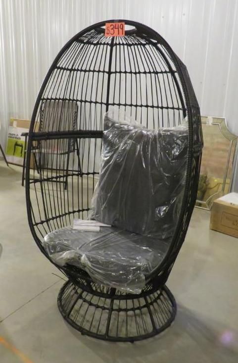 Black wicker egg-shaped chair with seat cushion, under plastic wrap, in a warehouse setting.