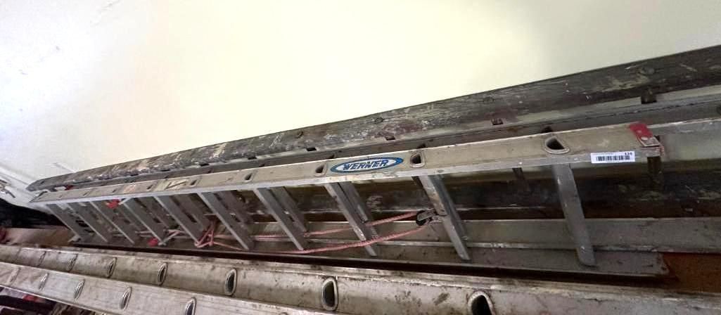 An aluminum ladder resting on a structure, against a white wall.