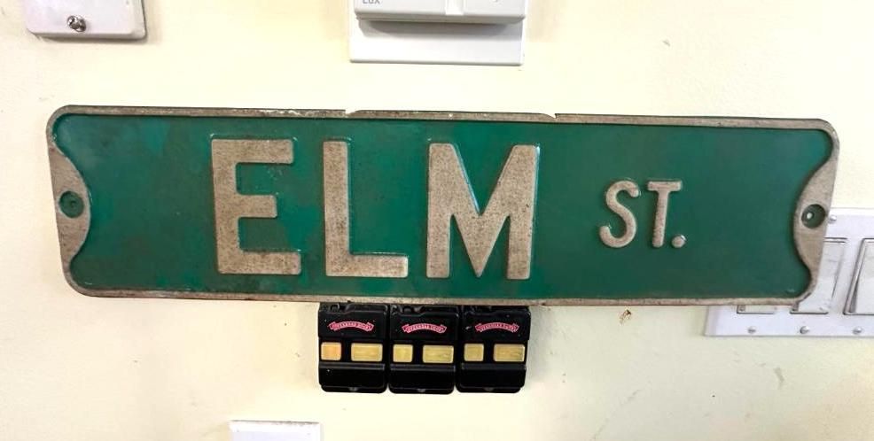 Green and silver Elm Street sign on a beige wall, with electrical breakers below and two light switches above.