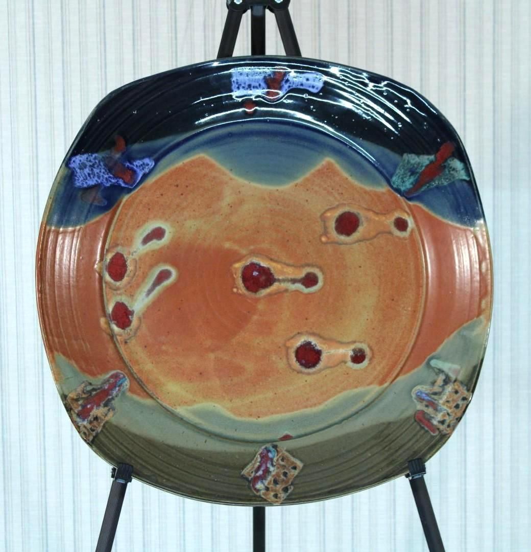 Ceramic plate with landscape design in orange, blue, and brown displayed on a black easel.