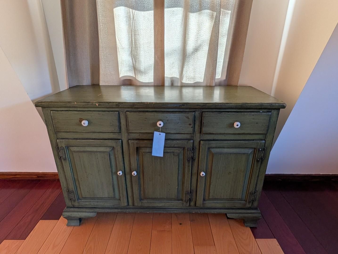 Green wooden sideboard cabinet with drawers and doors against a wall and curtain