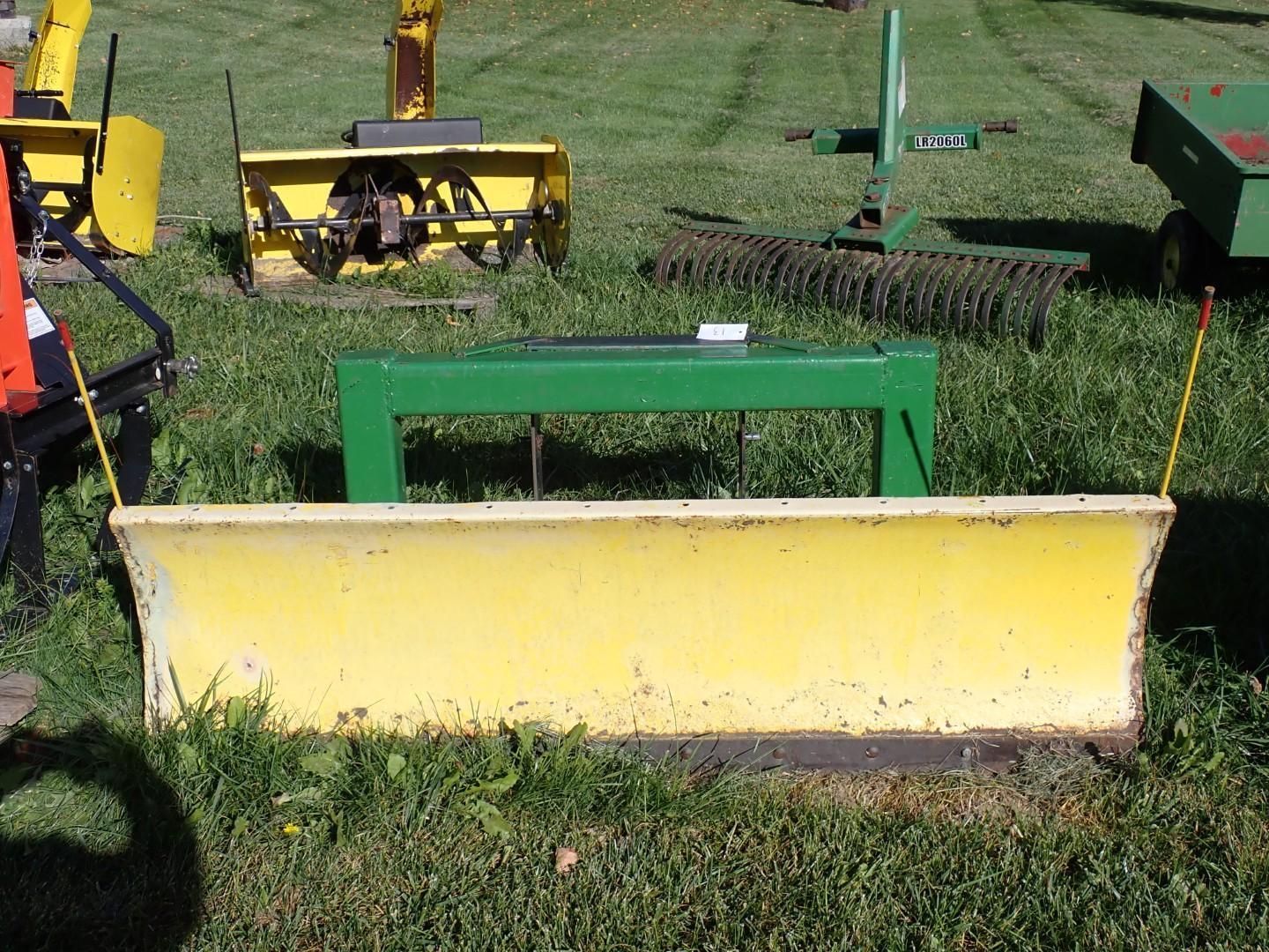 Yellow snow plow blade with green frame sitting on grass outdoors.