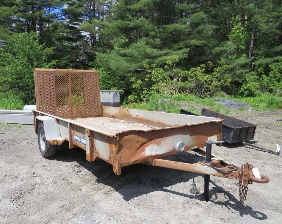 Metal utility trailer with a mesh front, outdoors on a gravel surface with trees in the background.