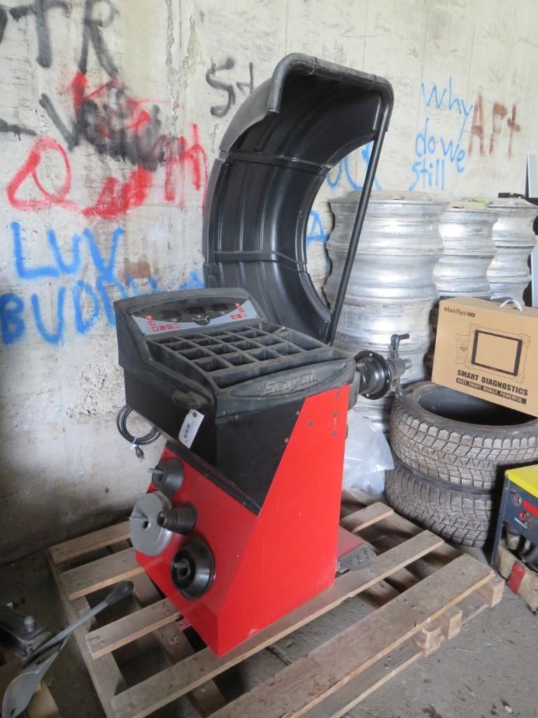 Red and black tire balancing machine on a wooden pallet, with tires and rims in the background.