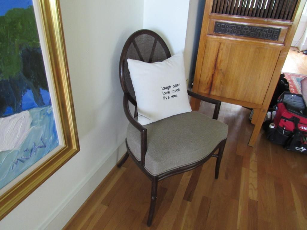 Chair with cushion, near a painting and wooden cabinet. Pillow has text, all on hardwood floor.