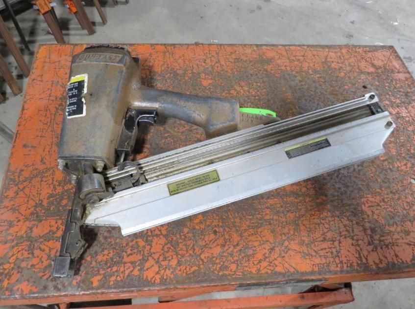 Pneumatic nail gun on an orange metal surface. Gray tool with a long magazine.
