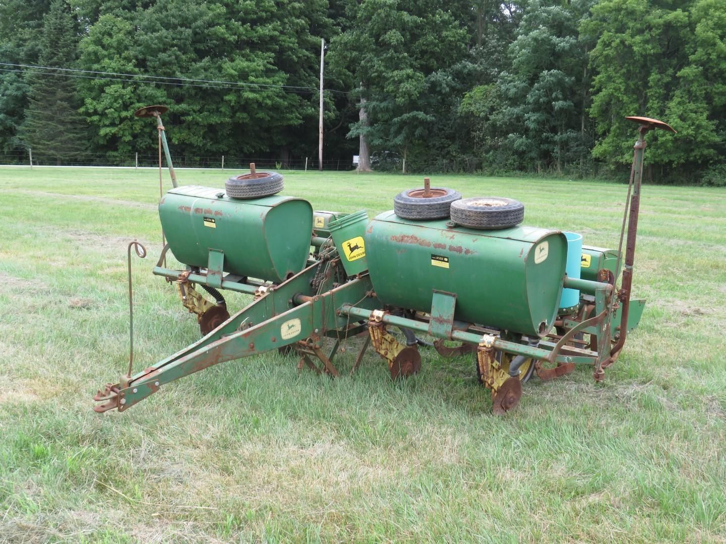 Green John Deere two-row planter on grass with trees in the background. Two tanks.