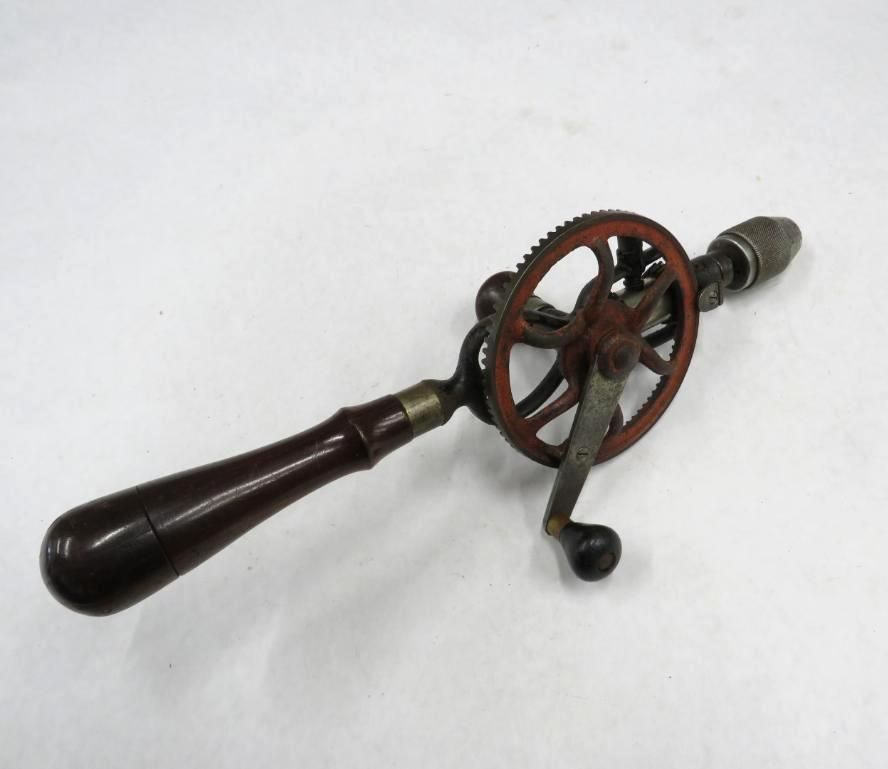 Hand-cranked drill with a wooden handle, metal gears, and a chuck for holding drill bits on a white surface.