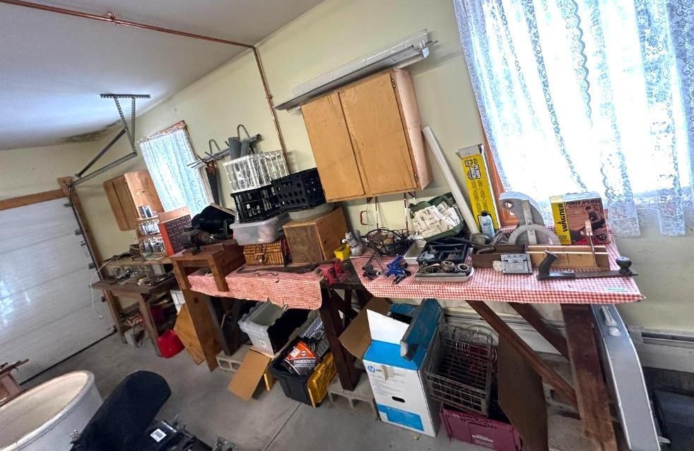 Garage interior with workbench covered in items, cabinets, and a window with lace curtain.