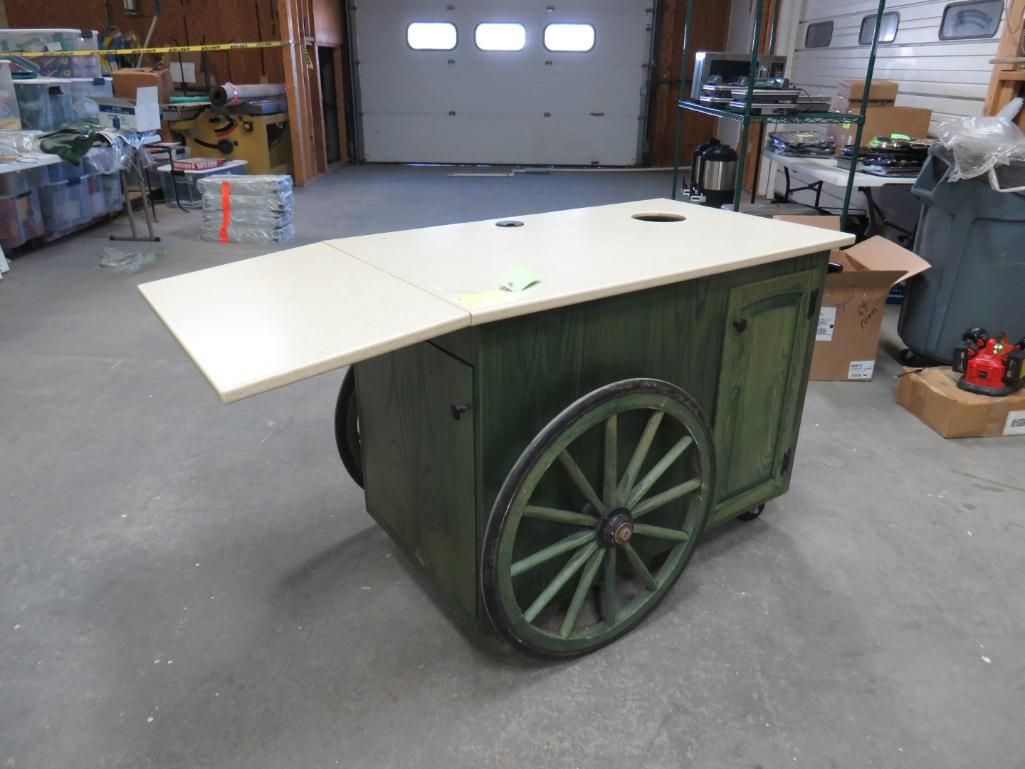 Green wooden cart with tabletop, a large wheel on each side, in a warehouse.
