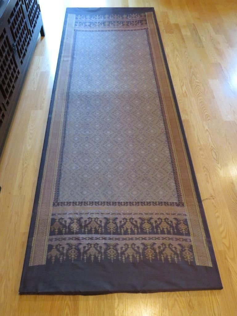 Long, rectangular, patterned rug on a wooden floor, with a brown border and dark center.
