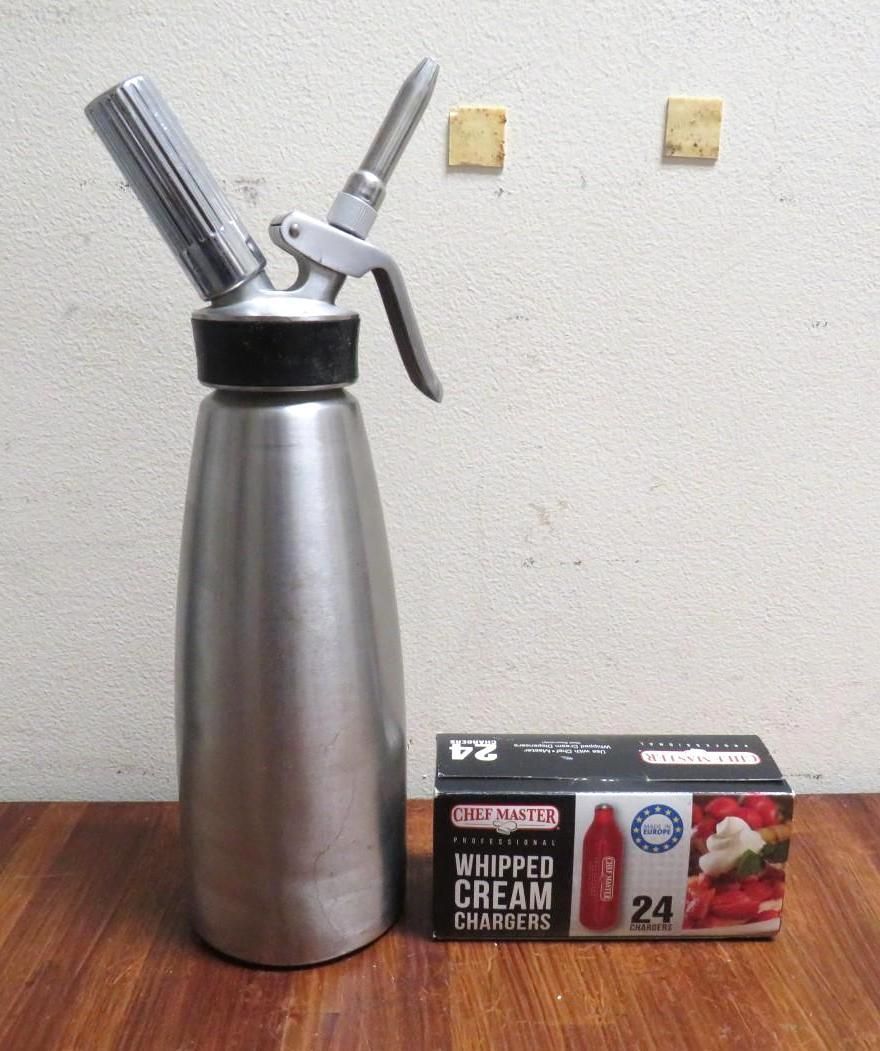 Whipped cream dispenser and cartridge box on a wooden surface against a plain wall.
