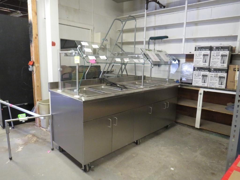 Stainless steel food buffet with glass sneeze guard in a storage room.