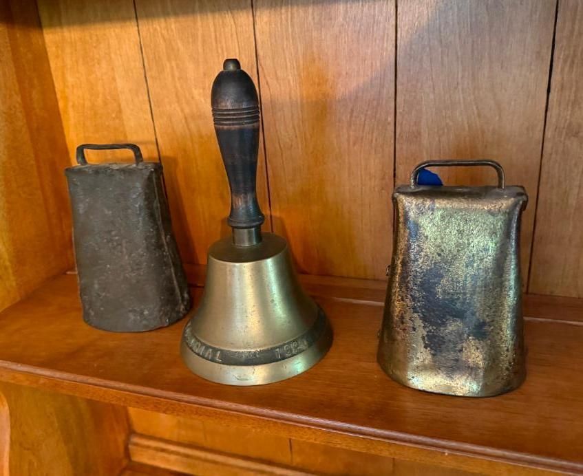 Three antique brass bells on a wooden shelf; one with a handle, two with rectangular loops.