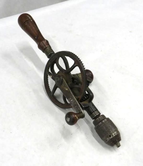 Hand-cranked drill with wooden handle and metal gears, lying on a white surface.