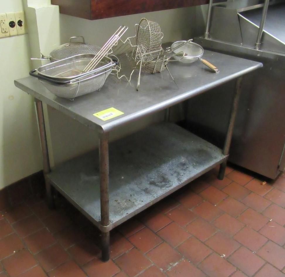 Stainless steel work table with shelf and cooking tools on top, in a commercial kitchen setting.