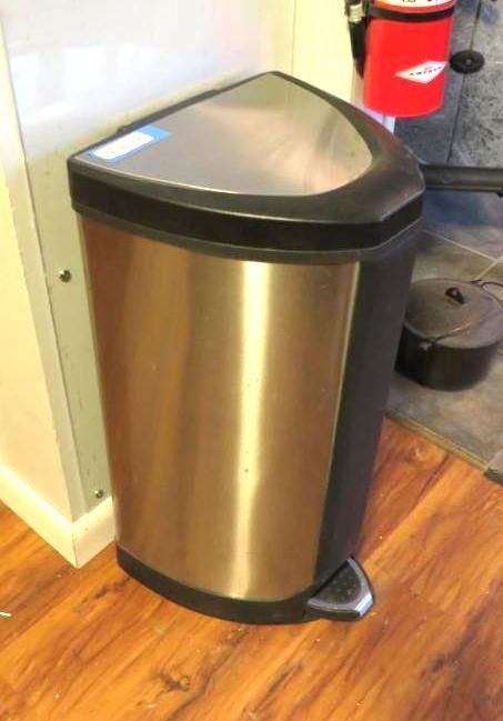 Stainless steel and black trash can with a foot pedal, on a wooden floor.