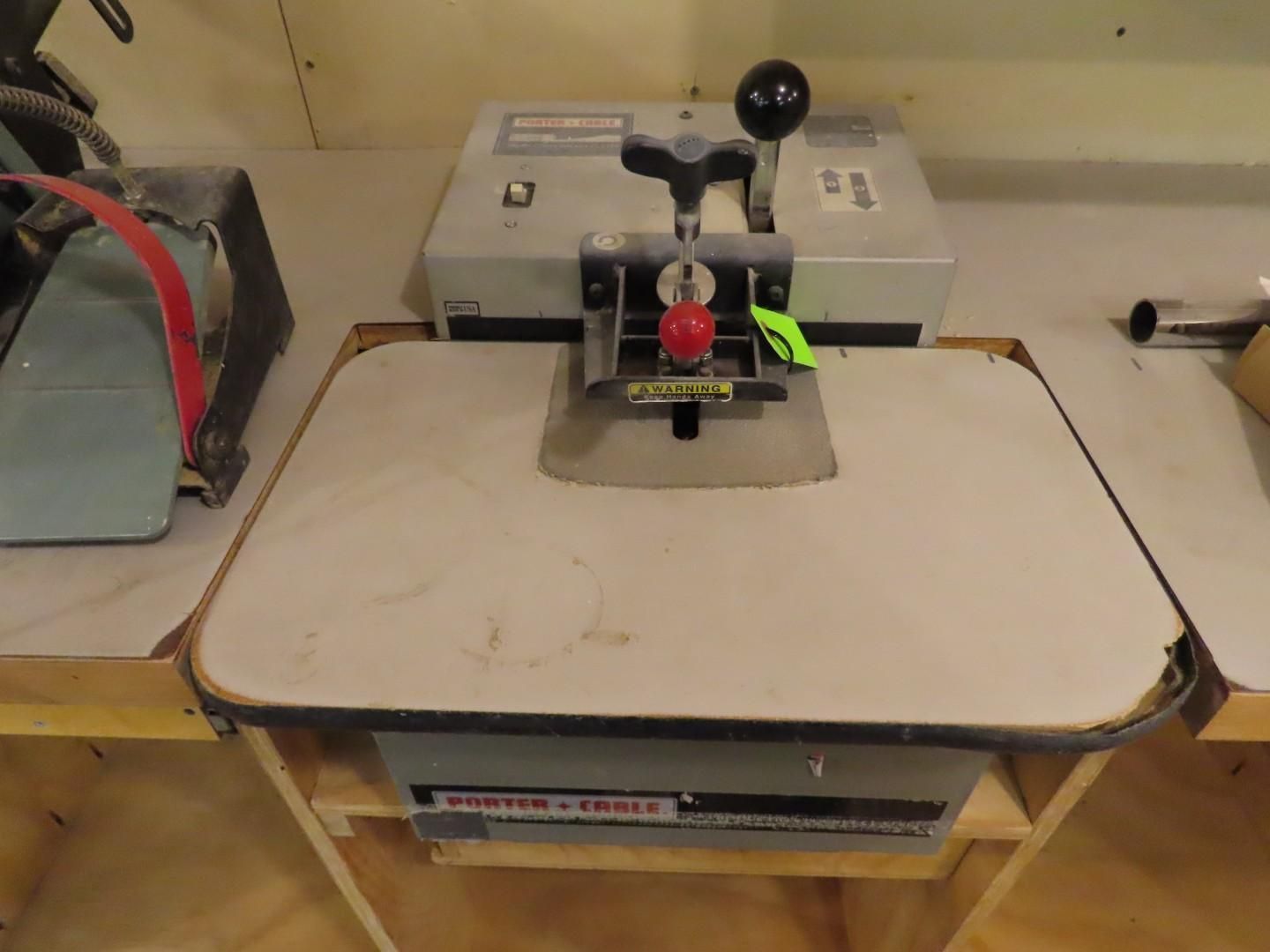 Small benchtop machine with black handles and a red knob on a worktable in a workshop.