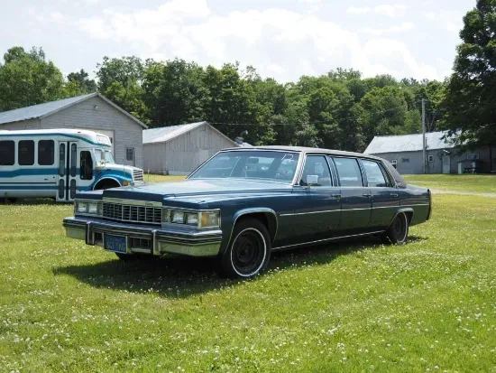 Dark blue limousine parked on grass near buildings and a bus. Sunny day.