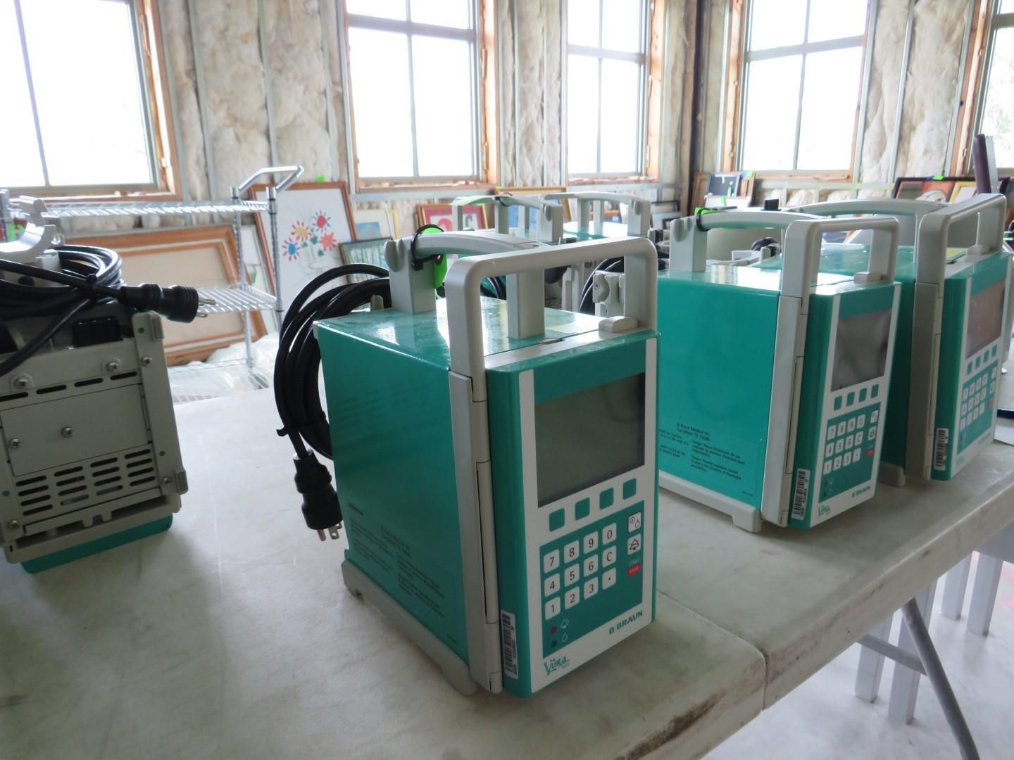 Several teal-colored medical infusion pumps lined up on a white table in a room with windows.