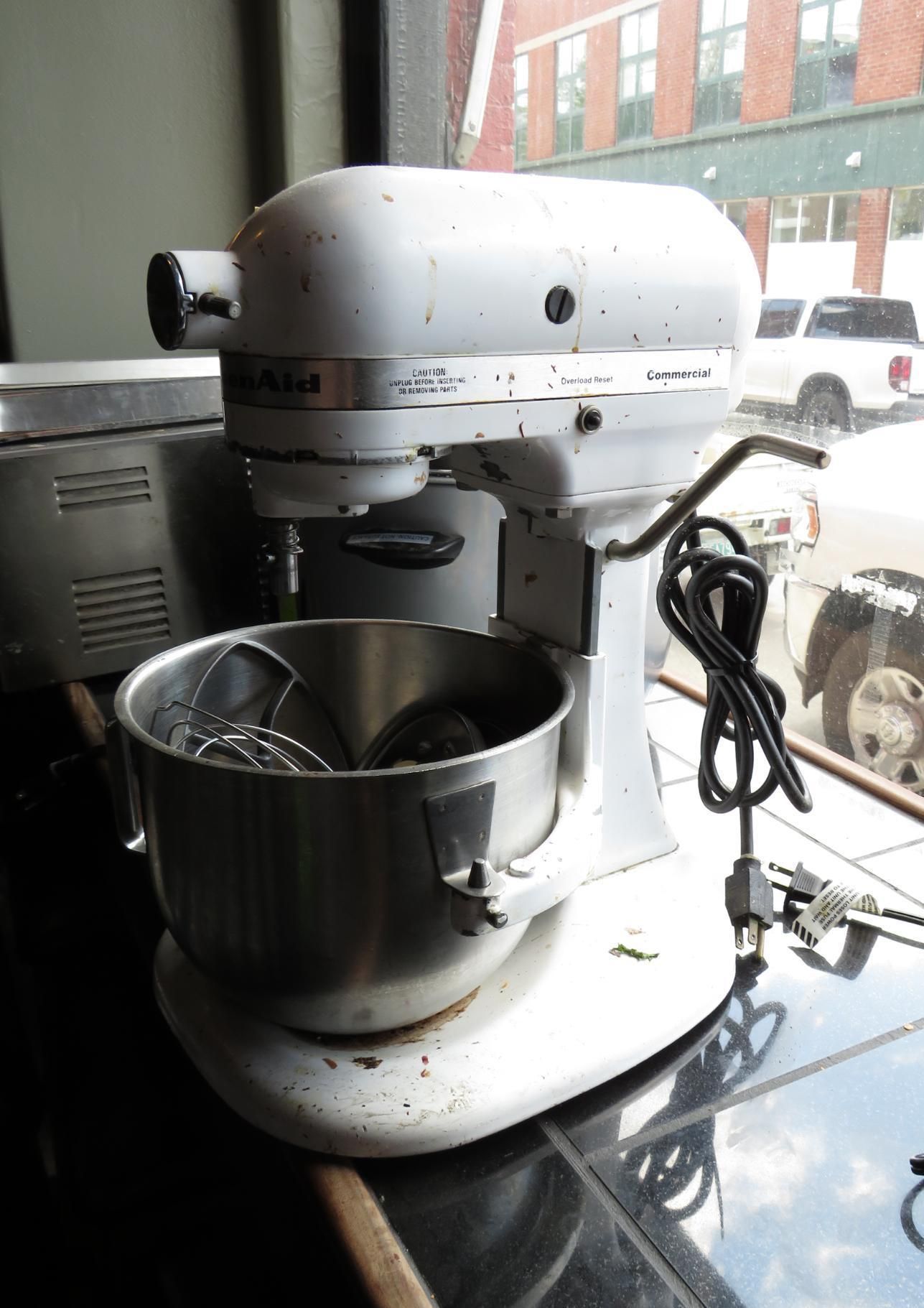 White KitchenAid stand mixer with a stainless steel bowl, by a window.