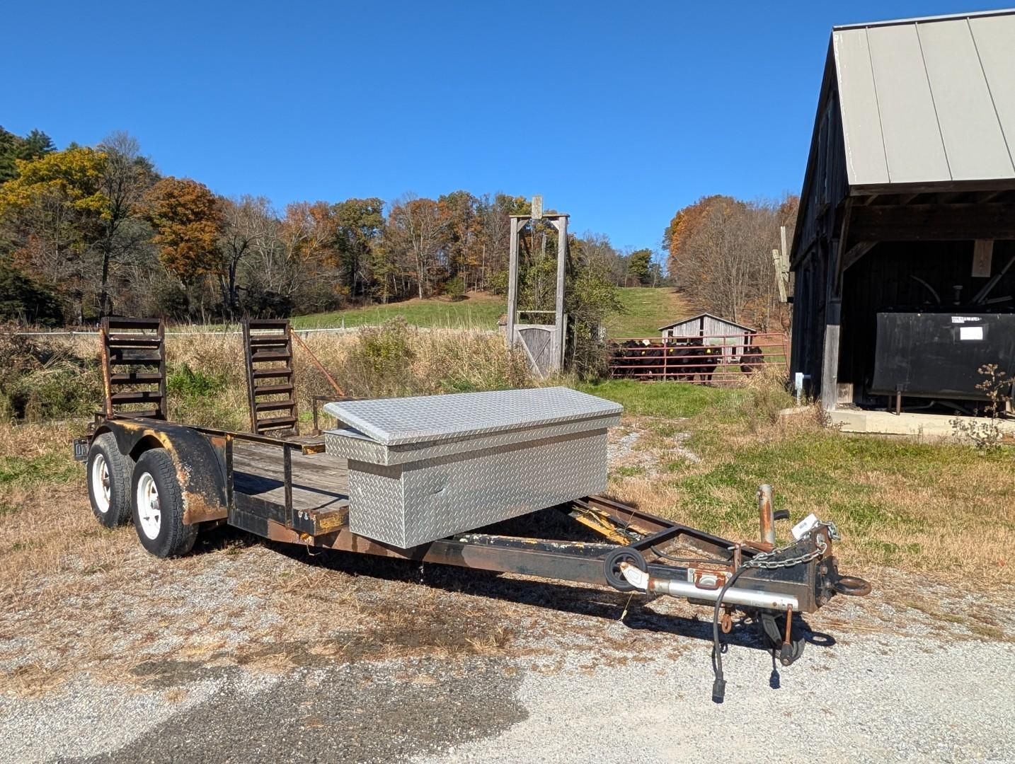 Utility trailer with a silver toolbox parked on gravel beside a barn on a rural property.