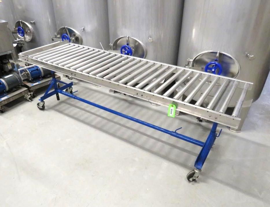 A long roller conveyor, silver and supported by a blue frame with wheels, in front of large silver tanks.