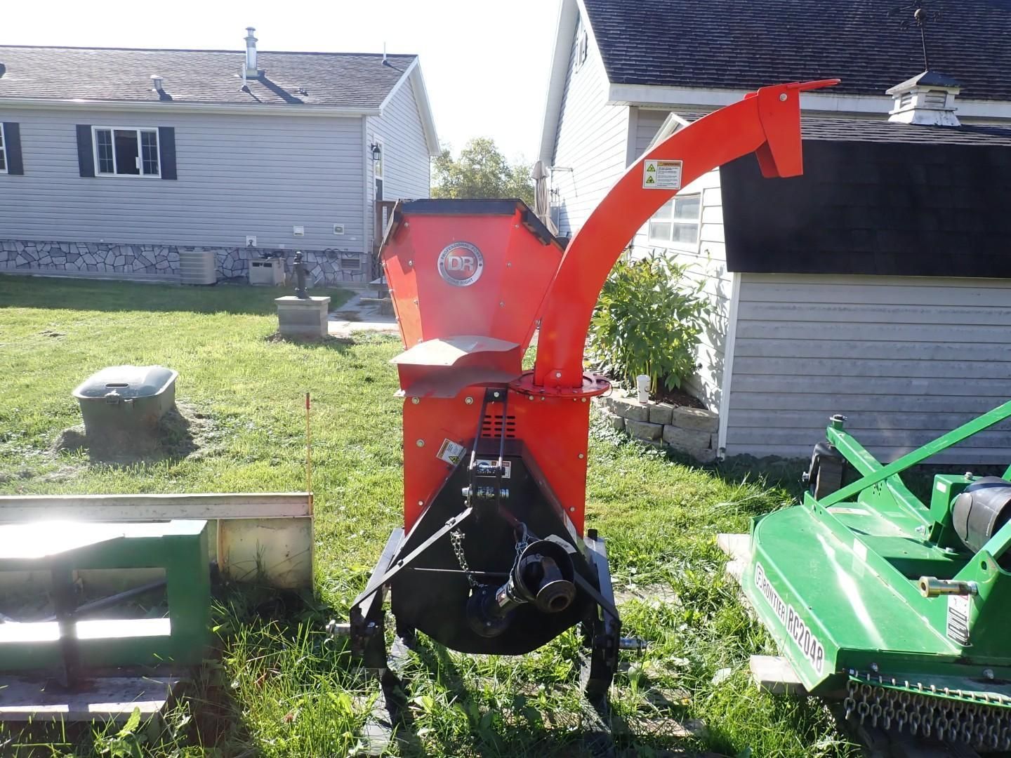 Red wood chipper with chute in a grassy yard beside houses, with a green attachment on the right