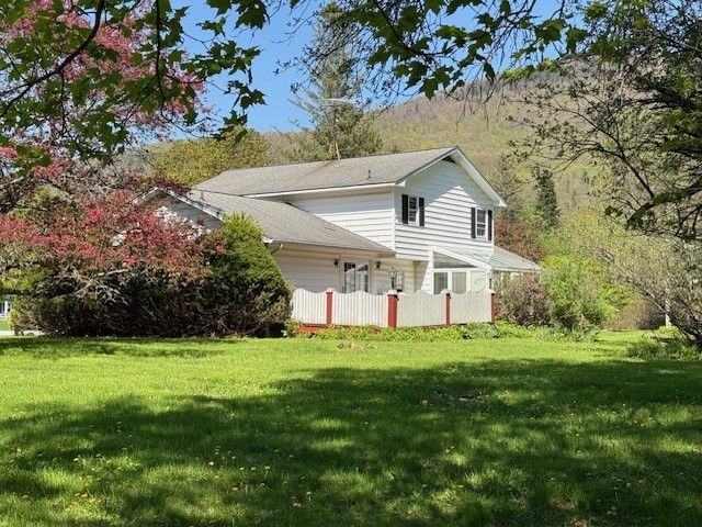 White two-story house with green lawn, trees, and mountain background on a sunny day.
