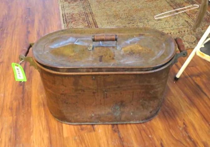 Copper storage container with wooden handles and lid, sitting on a wooden floor.